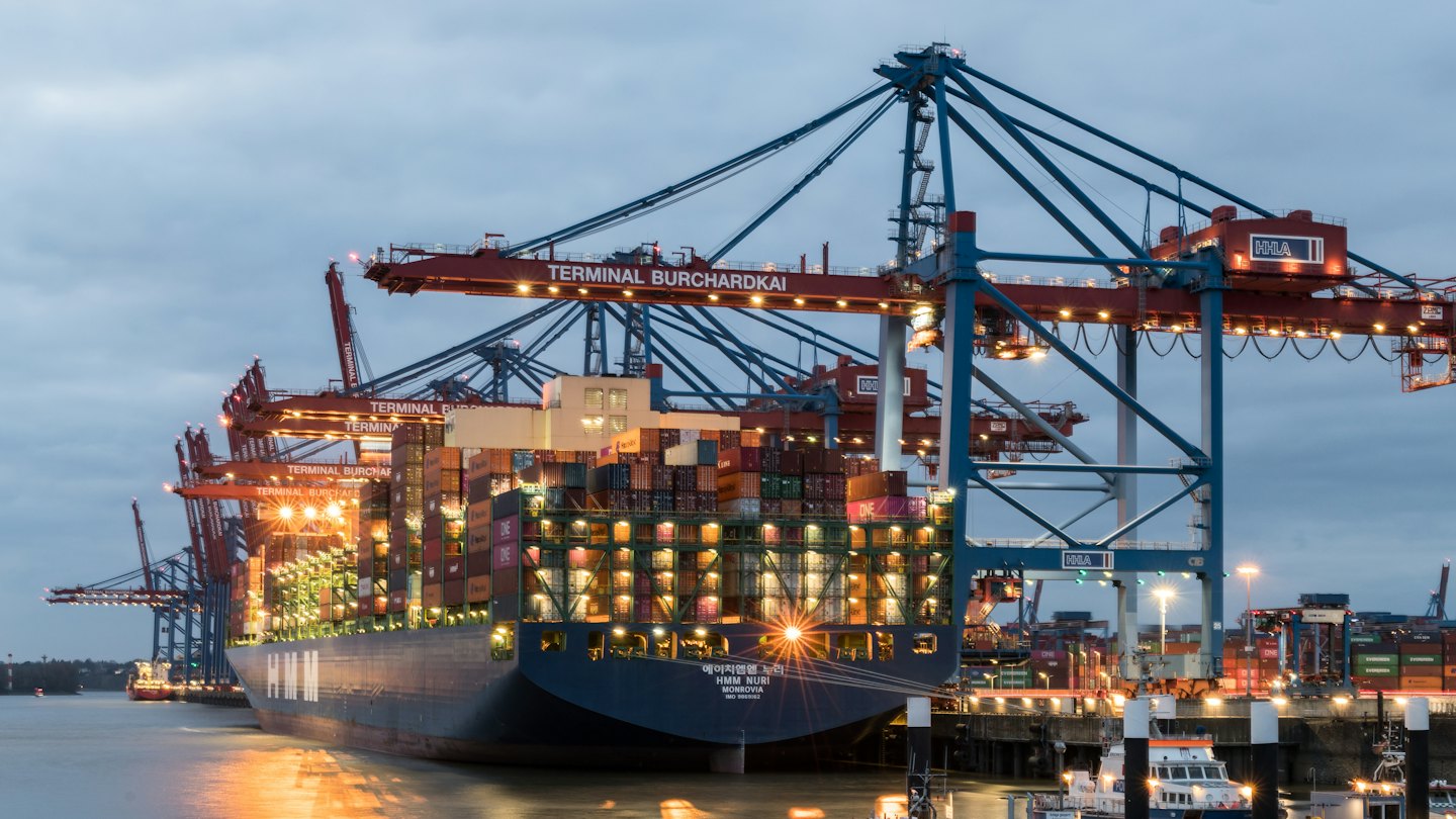 Container ship docked at port with gantry cranes at dusk.