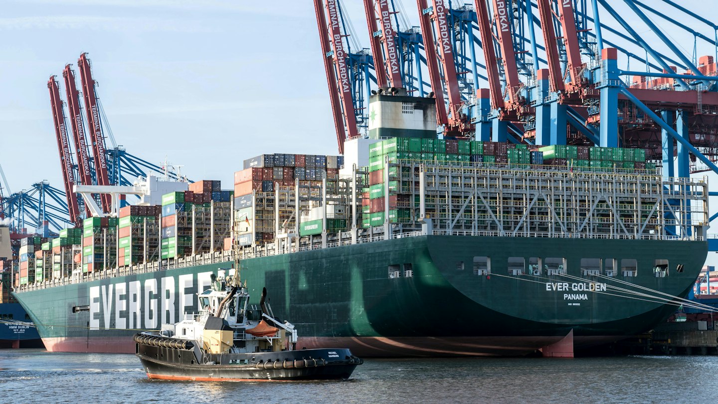 Container ship docked in bustling port with tugboat assistance.