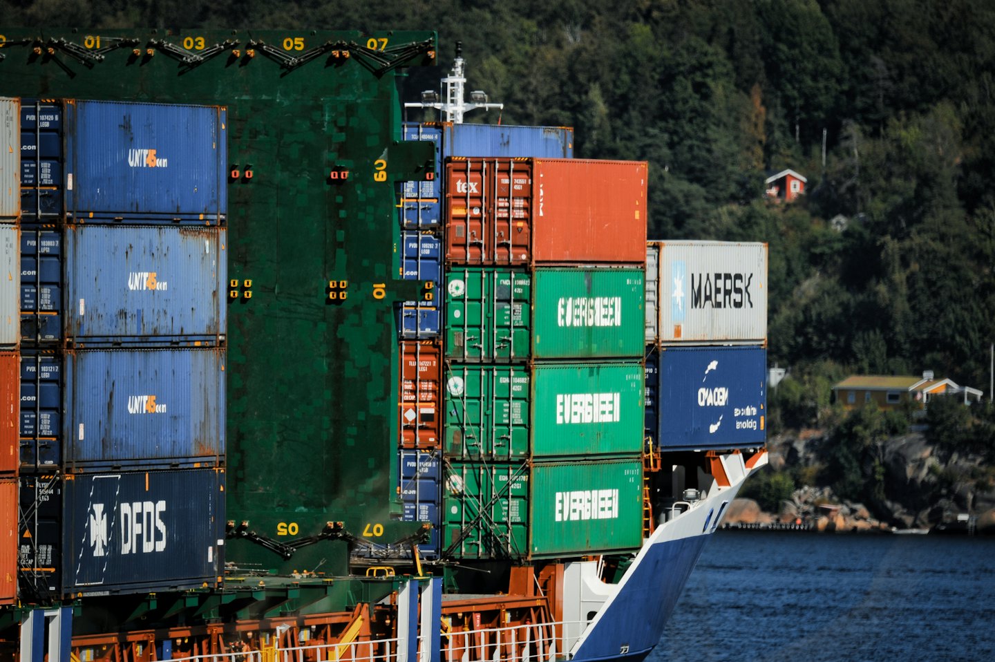 A large cargo ship sails past a forested hillside with houses.